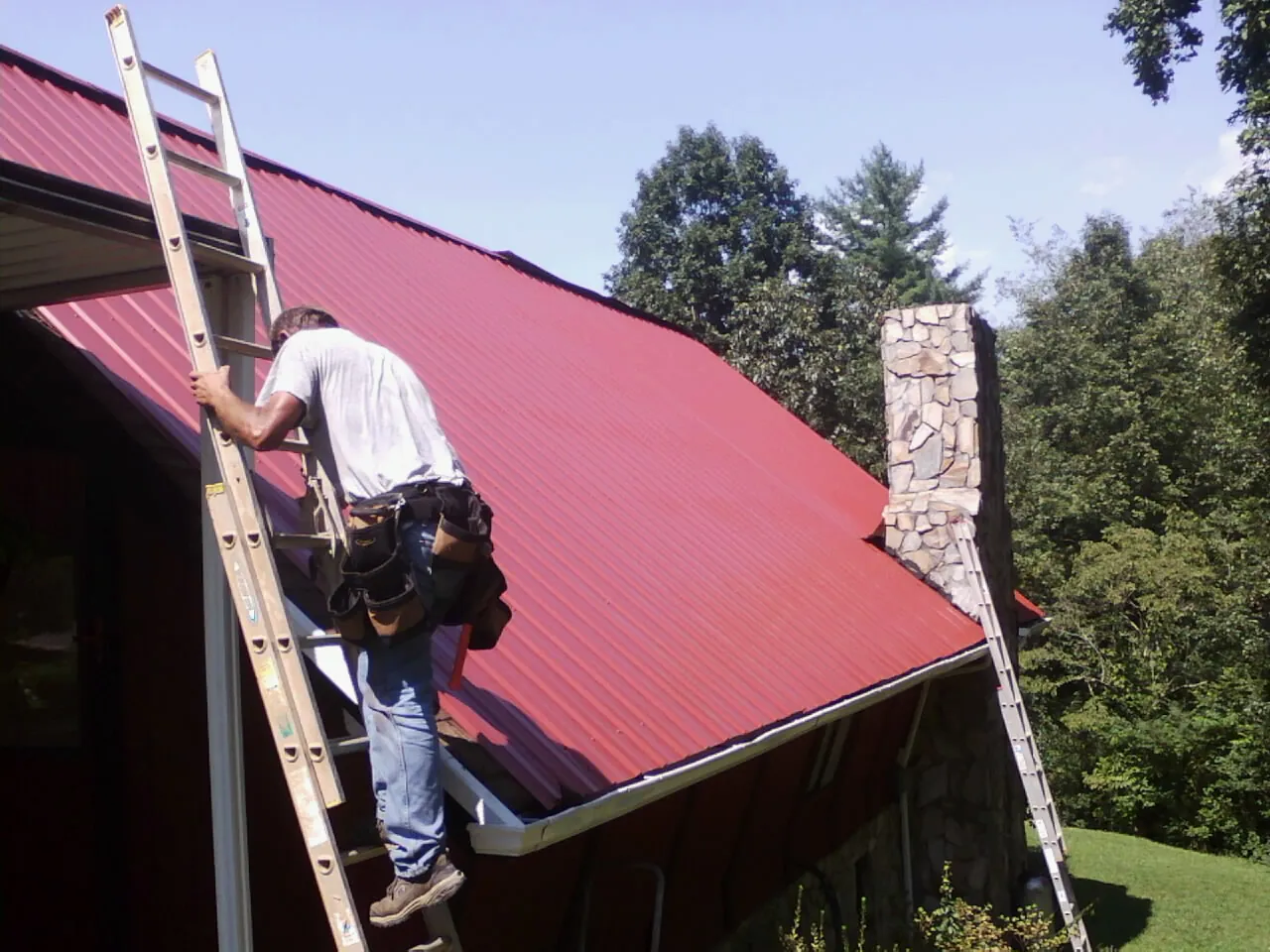 Roofer on a ladder beside a red metal roof and stone chimney — Soffit & Fascia Repair by Roofing Brothers in Boerne